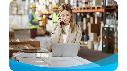 A smiling female manager in a business suit is talking on a phone and working on a laptop on top of wrapped pallets in a busy warehouse.